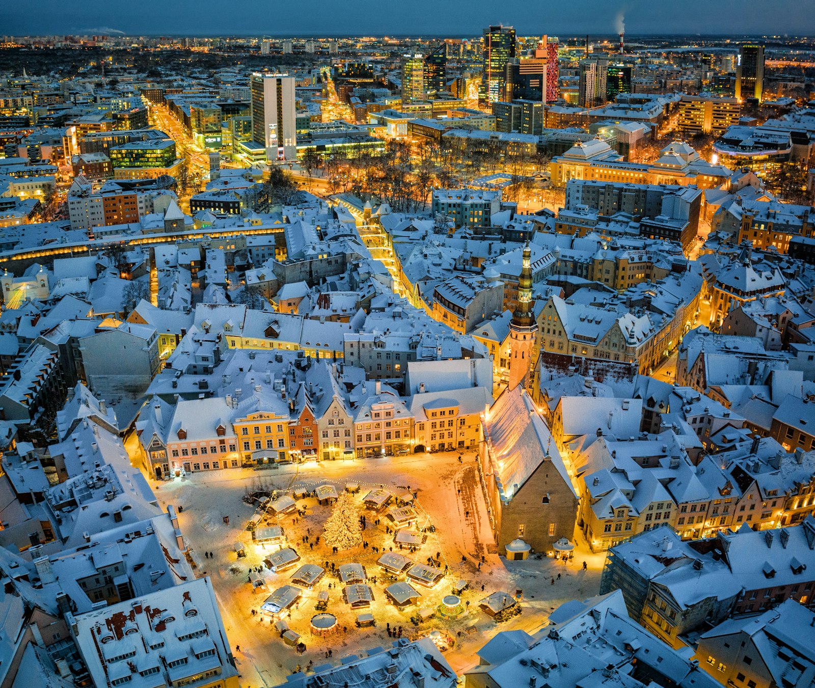 A cidade velha de Tallinn sob a neve à noite, a praça medieval iluminada pelo mercado de Natal, com o distrito comercial moderno atrás.